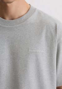 Jasnoszara bawełniana koszulka z okrągłym dekoltem, z subtelnym haftowanym logo na piersi w białej nitce. Gładka tekstura, standardowy krój.