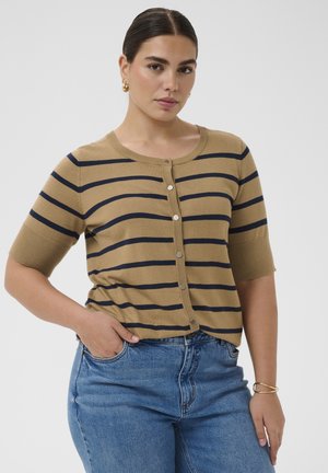 Sötét hajú nő kontyba fogott hajjal, arany karika fülbevalókkal, barna és fekete csíkos, gombos felsővel, kék farmernadrággal és arany karkötővel.