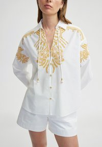 Camicia bianca con intricati ricami gialli, collo e scollatura a V. Presenta una chiusura frontale con bottoni e una vestibilità comoda. Abbinata a pantaloni corti bianchi.