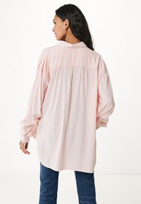 Blusa oversize rosa chiaro con maniche lunghe, dettaglio arricciato sul retro e colletto, abbinata a jeans scuri. Texture del tessuto liscia.
