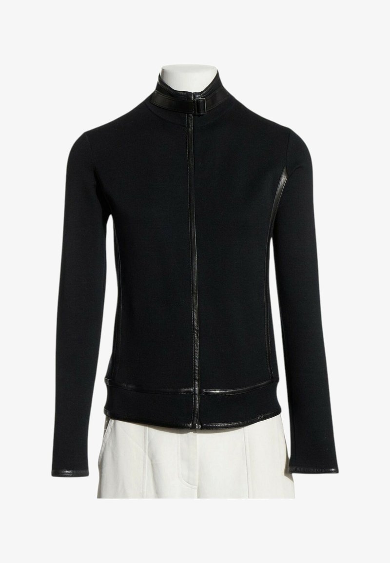 Veste noire cintrée à manches longues avec fermeture éclair frontale et col haut, associée à un pantalon blanc sur un mannequin sans tête.