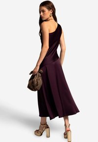 Robe violette à une épaule en tissu lisse, avec un ourlet évasé. Portée avec des sandales compensées dorées épaisses et un sac marron à motifs.