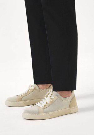 Béžové nízké pletené tenisky s bílými tkaničkami nošené s černými cropped kalhotami na bílém pozadí.