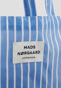 Niebieska torba na ramię w paski z błyszczącym wykończeniem, z białymi pionowymi paskami. Zawiera metkę z tkaniny z napisem "MADS NØRGAARD COPENHAGEN".