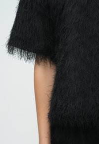 Czarny, fakturowany top z miękkim, ozdobnym wykończeniem z frędzlami. Krótkie rękawy rozszerzają się do szerokiej, pudełkowej sylwetki. Tkanina wydaje się miękka i warstwowa.