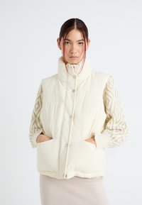 Veste rembourrée crème avec un col haut, dotée de manches en tricot décoratif à motif beige et d'un ourlet avec cordon de serrage. Boutons pression sur le devant et poches latérales.