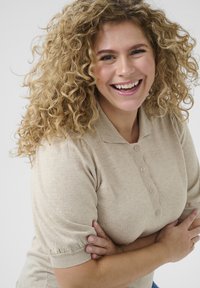 Beige polo-shirt met korte mouwen, een knoopsluiting, textuurstof en gathered cuffs. Het model heeft krullend haar en glimlacht.