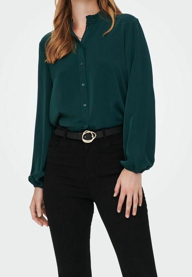 Femme portant un chemisier vert foncé boutonné avec des poignets froncés, rentré dans un pantalon noir taille haute avec une ceinture noire et une boucle dorée.