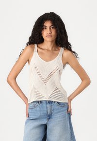 Knit tank top in beige with a chevron pattern, featuring wide straps and an open weave design, paired with loose blue jeans.