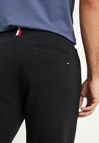 Czarne bawełniane spodnie z tylną kieszenią i małym logo w kolorach czerwonym, białym i niebieskim przy pasku. Gładka faktura, casualowy design.