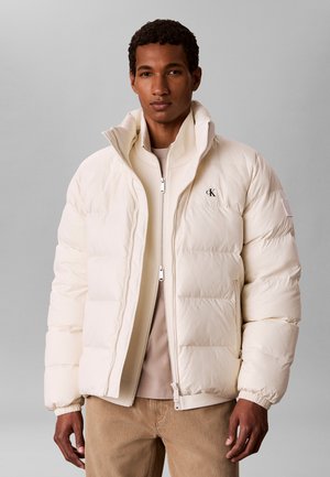 Man met een crèmekleurige donsjas en beige broek die staat tegen een effen lichtgrijze achtergrond.