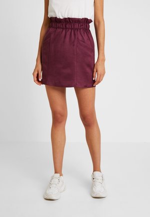 Femme portant une mini-jupe prune taille haute avec des poches à l'avant et des baskets blanches, debout devant un fond blanc uni.