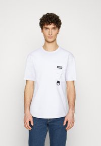 T-shirt bianca in cotone con scollatura rotonda, maniche corte e taschino sul petto con etichetta nera e attacco a loop nero.