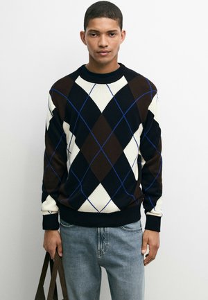Homme portant un pull à losanges noir, marron et blanc et un jean bleu clair, tenant un sac marron foncé sur un fond uni.