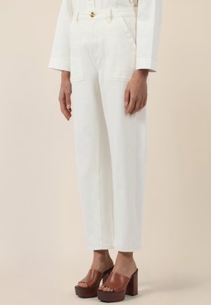 Kvinde iført hvide bukser med høj talje og forlommer samt brune plateauhøjhælede sandaler med åbne tæer på beige baggrund.