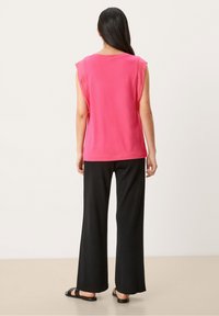 Růžový bezramínkový top s kulatým výstřihem, v kombinaci s širokými černými kalhotami a černými sandály. Tkanina vypadá měkce a lehce.