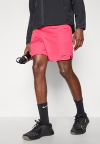 Vaaleanpunaiset Nike-urheilusortsit, joissa on kapea sovitus ja logo helman kohdalla. Yhdistetty mustiin sukkiin ja mustiin Nike-tossuihin, pitäen vesipulloa.