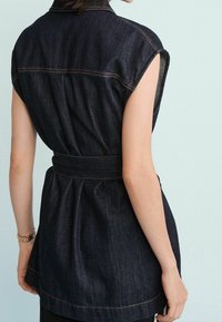 Ciemnoniebieska sukienka koszulowa z dżinsu z krótkimi rękawami, taliowana paskiem i plisami z tyłu. Charakteryzuje się kontrastowymi przeszyciami i usztywnionym kołnierzem.