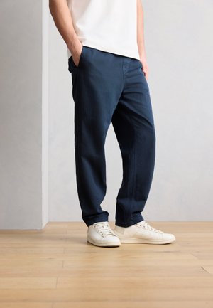 Άτομο που φοράει σκούρο μπλε casual παντελόνια και λευκά αθλητικά παπούτσια, στέκεται σε ανοιχτό ξύλινο πάτωμα με τα χέρια στις τσέπες.