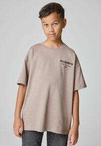 Beidzot apģērbs - bežs pārāk liels kokvilnas T-krekls ar īsām piedurknēm, kurā uz kreisā krūšu daļas ir melns teksts "ALLSAINTS" un papildus detaļas.