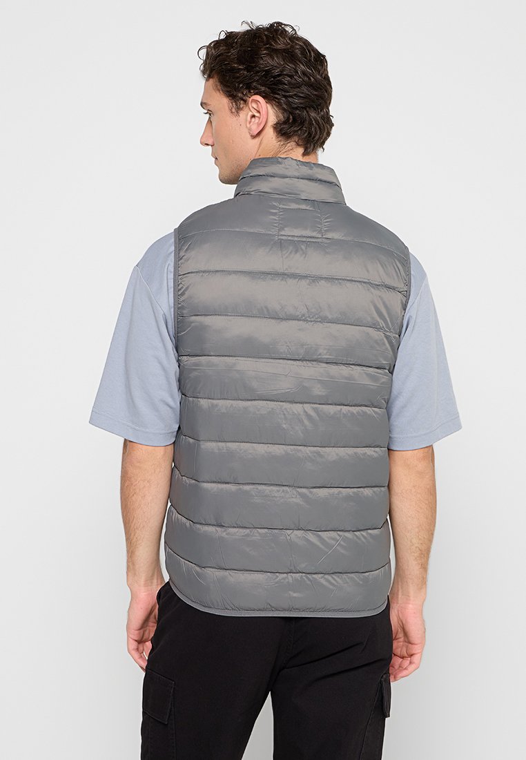 Chaleco acolchado gris con acolchado horizontal, cuello alto y una textura suave, llevado sobre una camisa de manga corta azul claro.