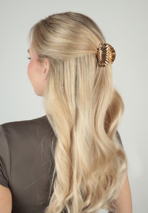 Donna bionda con capelli ondulati raccolti a metà, fissati da una lucida molletta dorata a forma di conchiglia, che indossa una maglietta scura a maniche corte, rivolta verso sinistra.