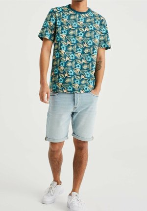 Camisa de manga corta en color teal con un patrón floral tropical, combinada con pantalones cortos de mezclilla azul claro con los puños doblados y zapatillas blancas.
