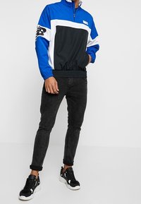 Blå och svart zip-up jacka med elastisk nederkant, vita accenter och logotyp; kombinerat med svarta slim-fit jeans och svarta sneakers.