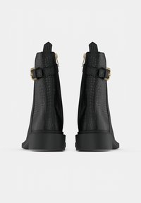 Pár černých kotníkových bot s krokodýlím vzorem, zlatými přezkami a bočními zipy, pohled zezadu na bílém pozadí.