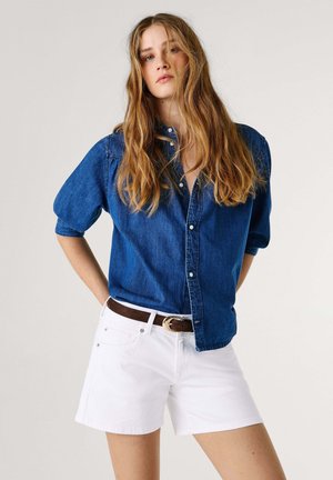 Giovane donna con capelli lunghi che indossa una camicia di jeans blu e pantaloncini bianchi con una cintura marrone, in piedi contro uno sfondo semplice.