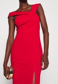 Kobieta w czerwonej sukience bez ramiączek z rozcięciem do uda, trzymająca w lewej ręce małą błyszczącą złotą kopertówkę.