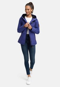 Lila vattentät jacka med huva, dragkedja och läderdetaljer, kombinerad med mörka jeans och vita sneakers. Slät textur, modern design.