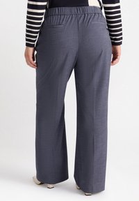 Breda byxor i mörkgrått, med en texturerad tyg, elastisk midjeband och två bakfickor. Sko med klossklack är synliga.