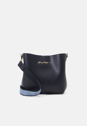 Borsa a tracolla in pelle blu navy con parte superiore curva, logo dorato e tracolla regolabile a strisce blu e bianche. Texture liscia, design minimale.