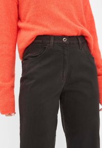 Jeans in cotone marrone con taglio dritto, cinque tasche e chiusura con bottone, abbinati a un maglione lavorato a maglia arancione. Tessuto strutturato visibile.
