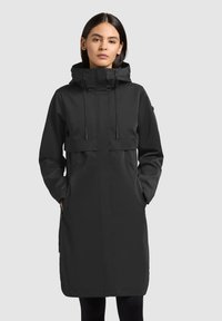 Ženska, oblečena v dolg črn dežni plašč s kapuco in sprednjimi žepi, stoji pred enobarvnim svetlo sivim ozadjem.