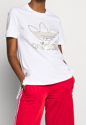 Osoba nosząca białą koszulkę Adidas z złotym logo oraz czerwone spodnie dresowe Adidas z białymi paskami, z rękami w kieszeniach, stojąca na tle gładkiej ściany.