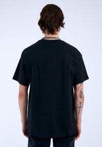 Camiseta de algodón negra con mangas cortas, cuello redondo y corte relajado. Tatuajes notables visibles en el brazo.