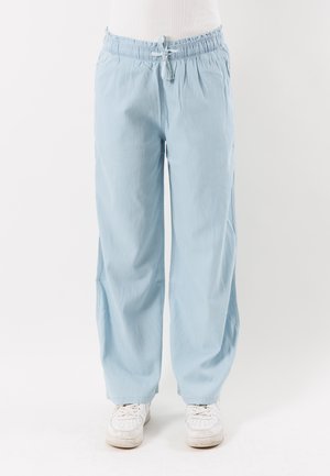 Pantaloni azzurri larghi con vita elastica e coulisse, indossati con sneakers bianche su sfondo bianco.