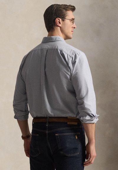 Homme avec des lunettes portant une chemise gris clair à boutons avec manches retroussées et un jean foncé, debout, dos principalement tourné vers la caméra.