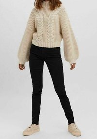 Gruby sweter z dzianiny w kolorze kremowym, z wysokim kołnierzem i wzorami w warkocze, w zestawieniu z czarnymi skinny jeansami i beżowymi sneakersami.