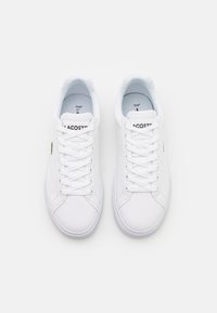 Hvite lerrets sneakers med snøring og rund tå. Har Lacoste-merke og logo på siden. Glatt tekstur og lett design.