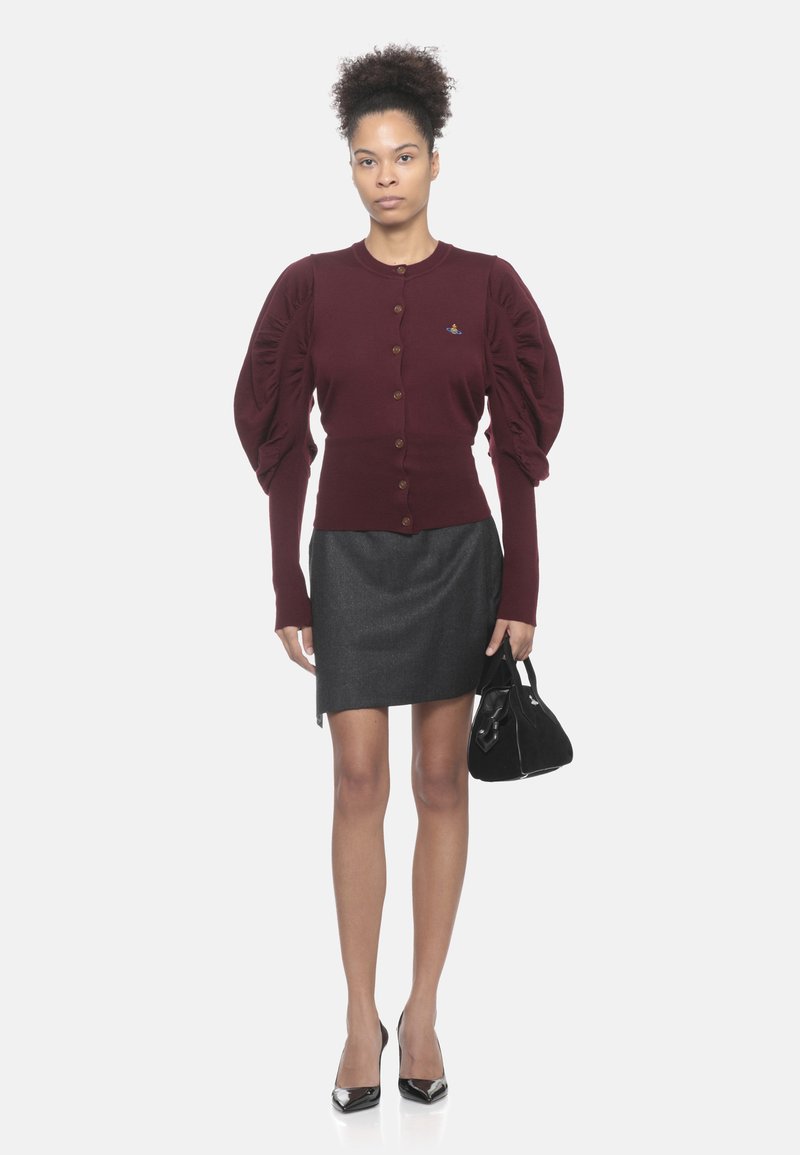 Donna che indossa un cardigan bordeaux con bottoni e maniche a sbuffo, gonna grigia scura, tacchi neri, con una piccola borsa nera in mano, in piedi su uno sfondo bianco.