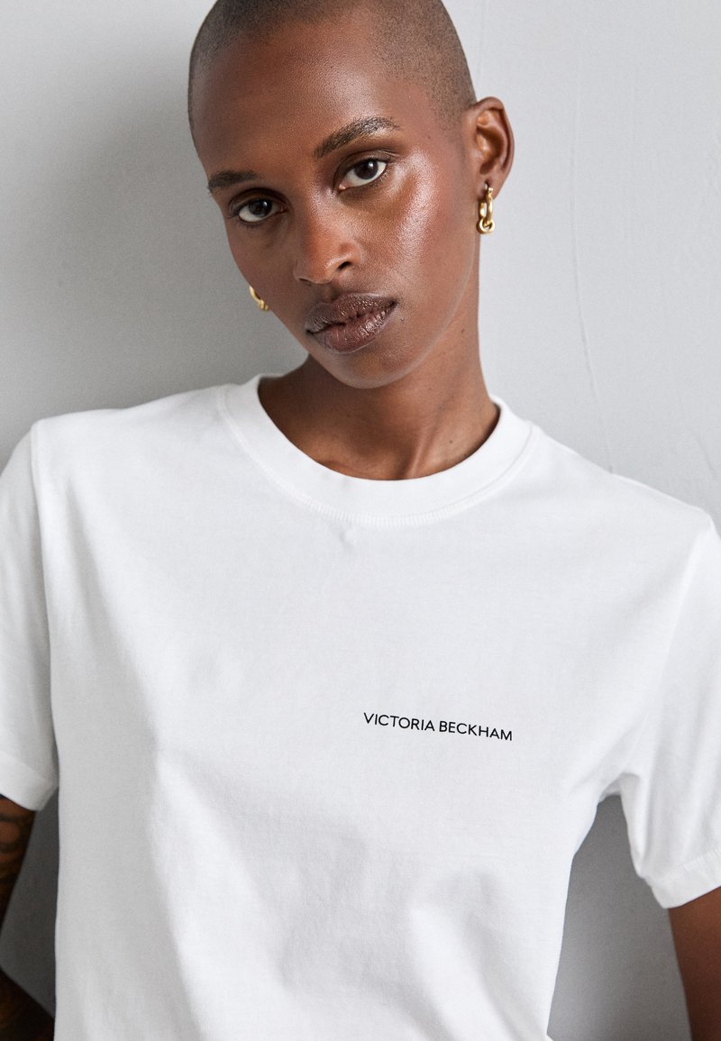 Camiseta de algodón blanca con cuello redondo y mangas cortas, con "VICTORIA BECKHAM" impreso en negro en el pecho. Textura suave.