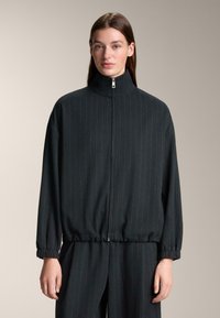 Fekete cipzáras dzseki magas gallérral, függőleges vékony csíkozással, rugalmas mandzsettákkal és laza szabással. Simára tapintó anyag.