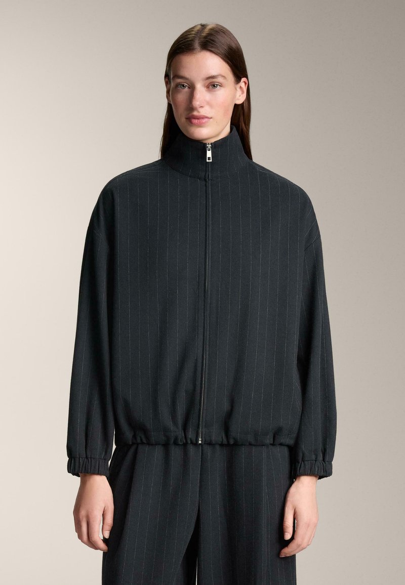 Fekete cipzáras dzseki magas gallérral, függőleges vékony csíkozással, rugalmas mandzsettákkal és laza szabással. Simára tapintó anyag.