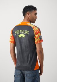 Kortærmet trøje med en mørkegrå krop, livlige orange og røde mønstrede ærmer samt et grafisk design på ryggen.