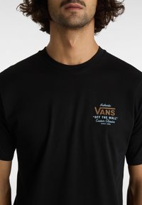 Černé bavlněné tričko s logem na hrudi v hnědé a modré barvě, které říká "Autentické VANS 'OFF THE WALL' Vlastní klasiky od roku 1966."