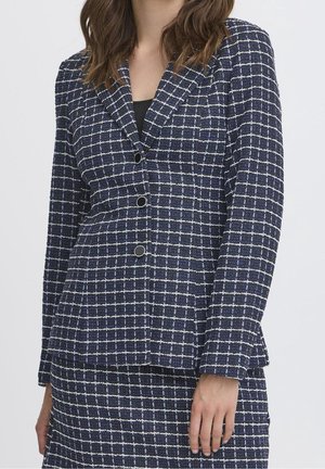 Kvinde iført en marineblå tekstureret blazer- og nederdelssæt med hvidt ternet mønster og sort top under, stående med den ene hånd afslappet.