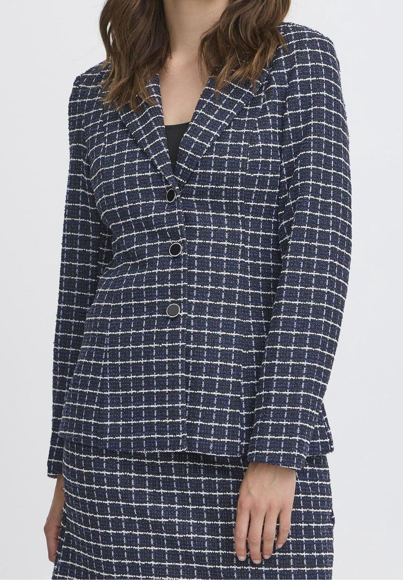 Femme portant un ensemble blazer texturé bleu marine et jupe avec motif quadrillé blanc et un haut noir en dessous, debout avec une main détendue.